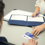 A white woman passes a business card across a desk to a white man.