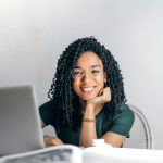A black woman smiles. She is working behind a laptop and her chin rests on her hand.